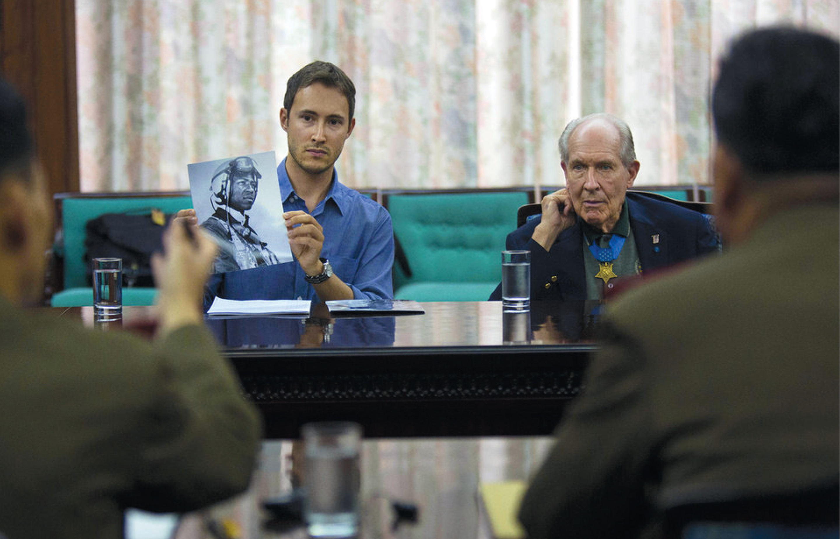 Adam Makos, left, author of Devotion: An Epic Story of Heroism, Friendship, and Sacrifice, holds up a photo of Jesse Brown in a meeting with officials in North Korea in 2013 while Thomas J. Hudner Jr., right, looks on.