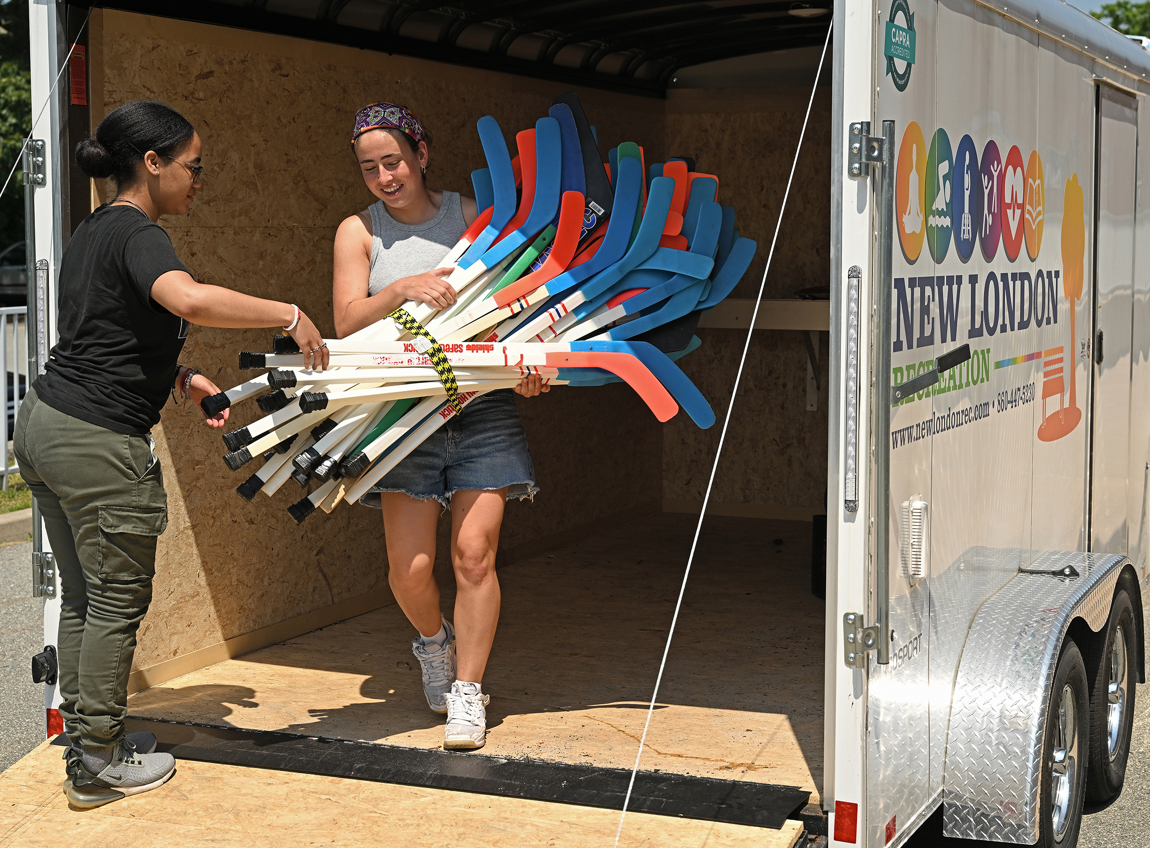 Two students struggle to carry a bundle of street hockey sticks out of a transport trailer