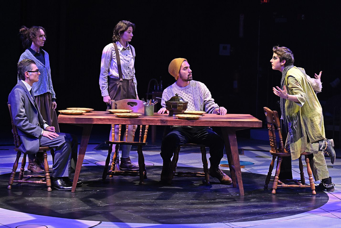 One actor gestures while speaking to the rest sitting around a table in a scene from a play in a black box theater.