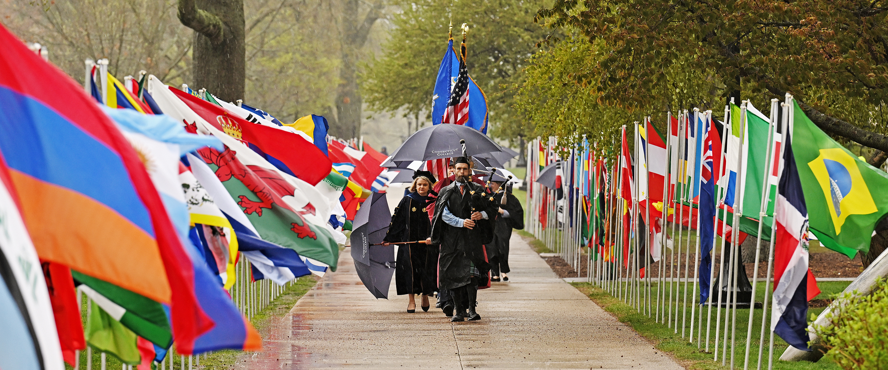 A procession of academics, led by a bagpiper and a color guard with U.S. and Connecticut flags, marches down a walkway flanked by international flags.