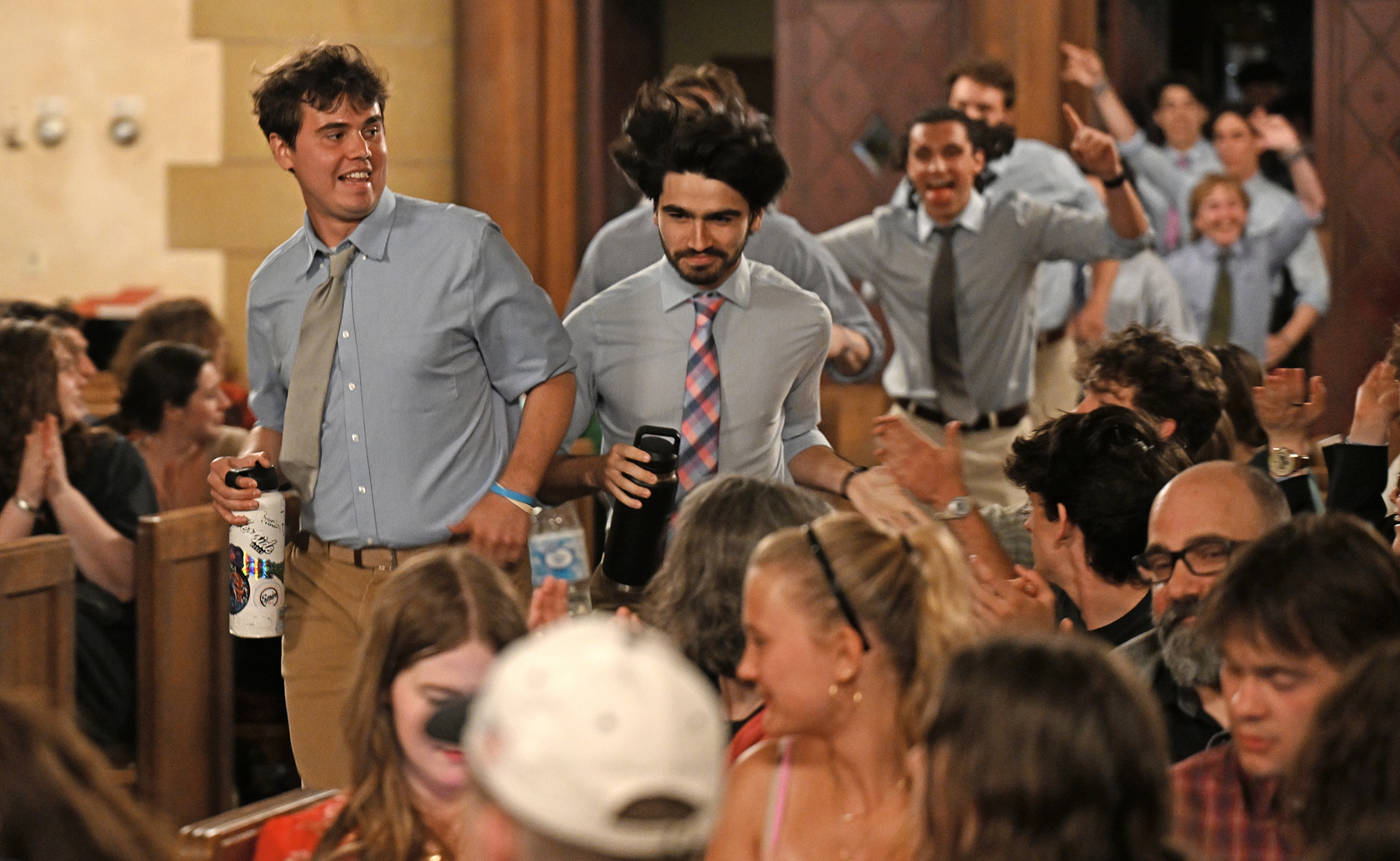 Students in matching blue shirts and non-matching ties run up the aisle of a chapel to take the stage for an a cappella concert.