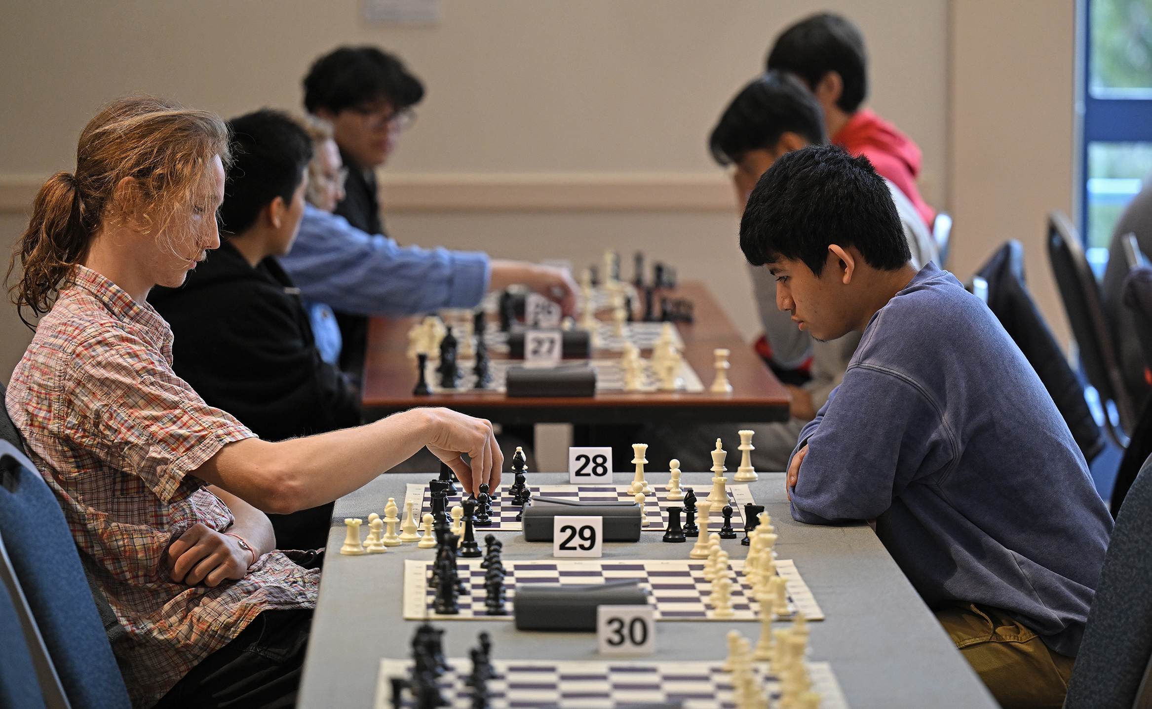 A college student with long blond hair plays chess in a crowded conference room during a tournament.
