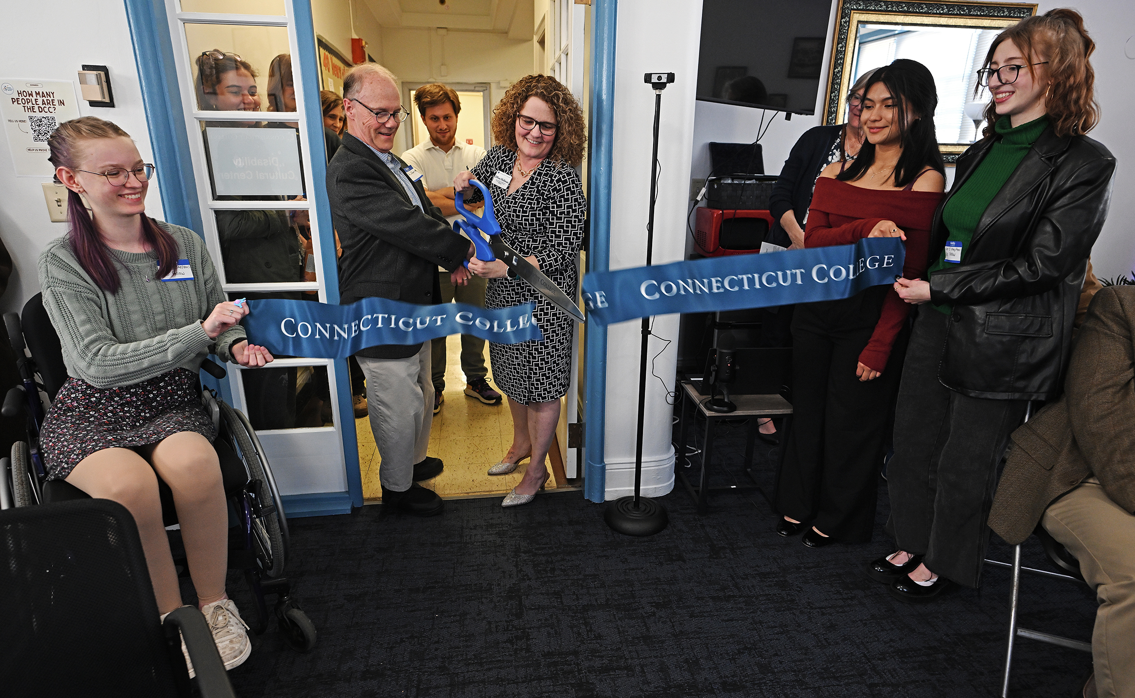 Students, including one in a wheelchair, hold a ribbon as two people with giant scissors cut the ribbon for the grand opening of a center for students with disabilities.