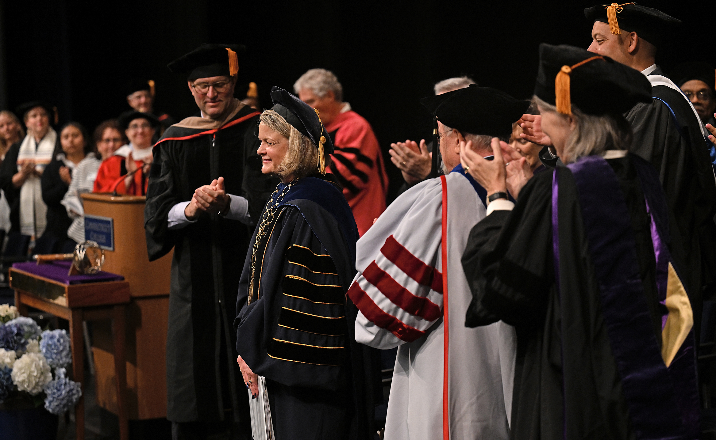 People in academic regalia stand and applaud at the inauguration of a new college president.