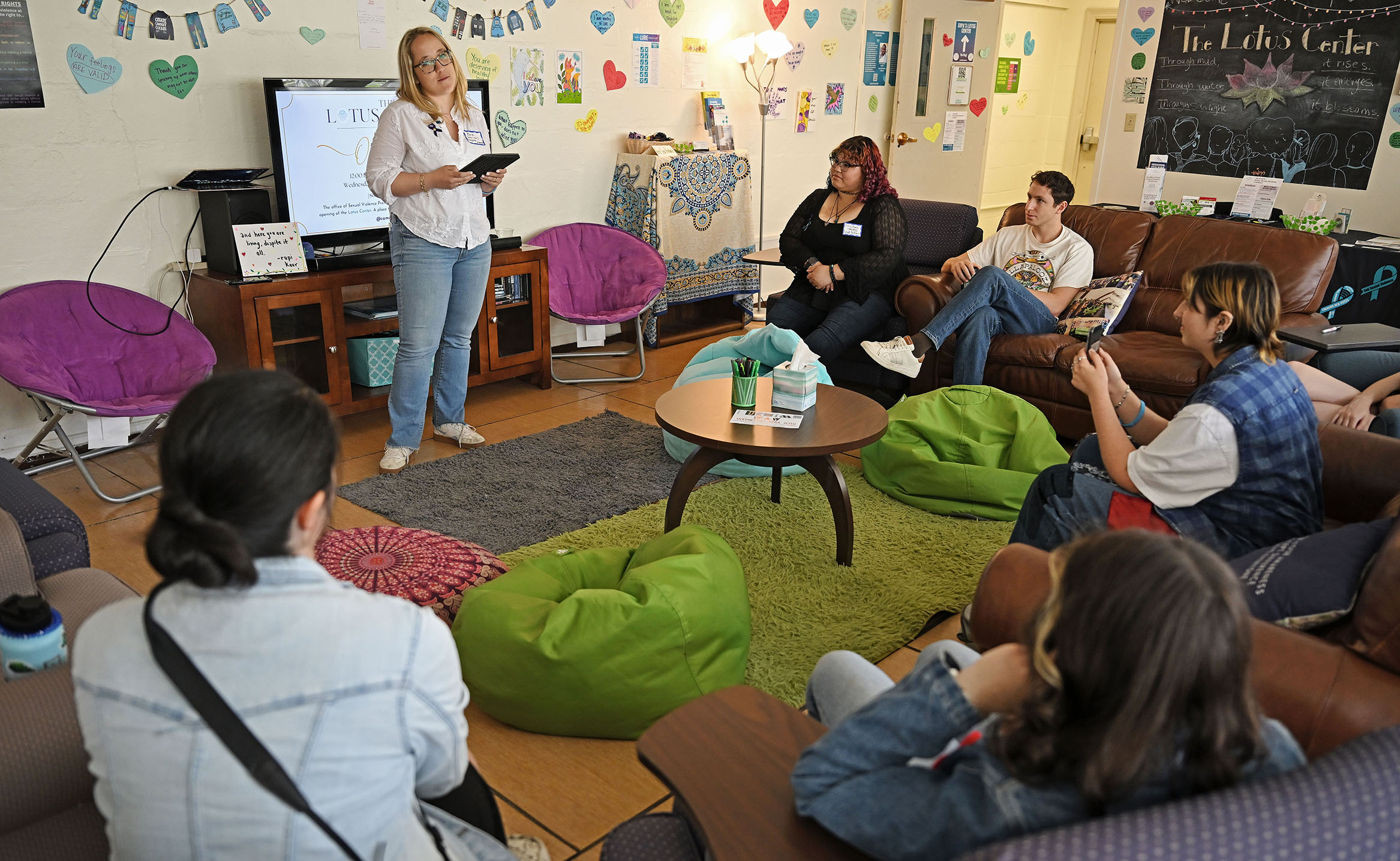 A speaker stands in front of a room full of casual and comfortable furniture to deliver a keynote address at a dedication of the space.