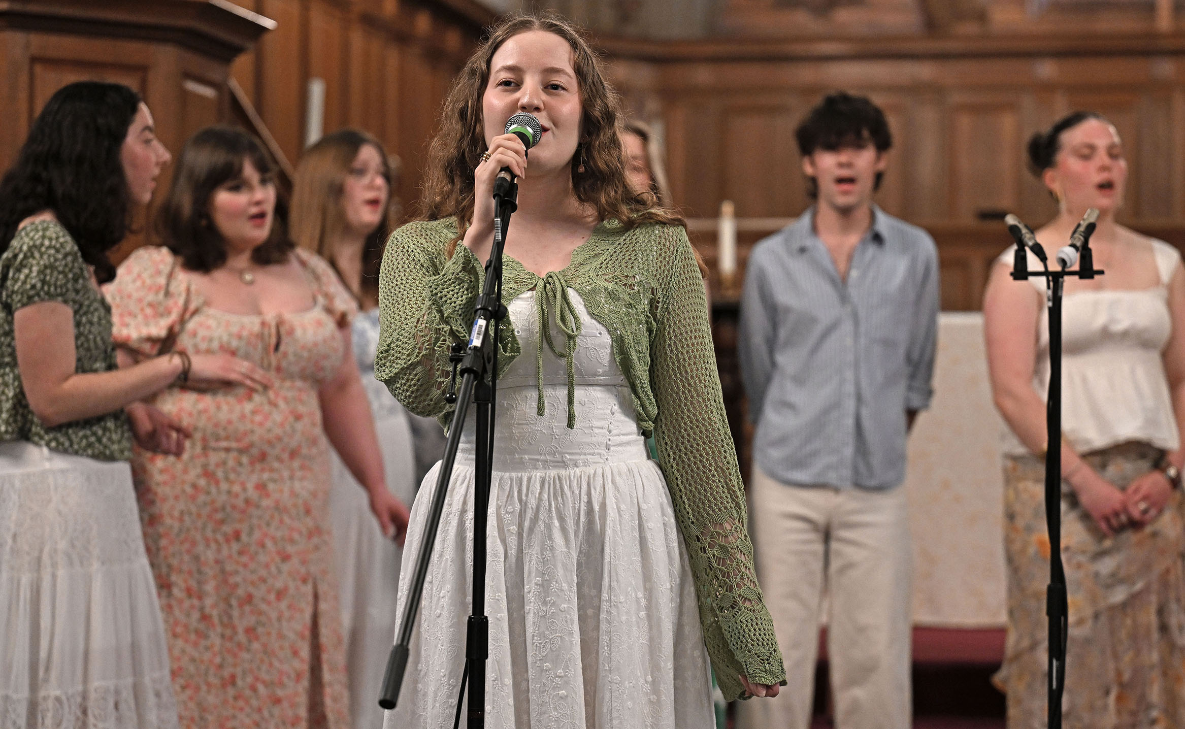 A female singer solos in front of an a cappella group performing in a college chapel.