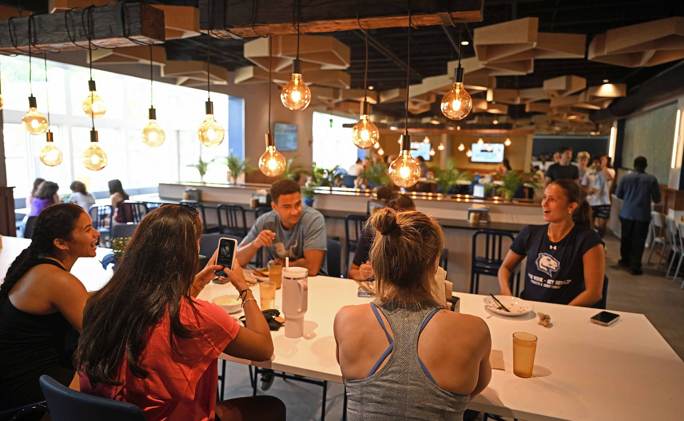 Students gather around a table in a college dining hall with bare bulb lighting and warm wood tones.