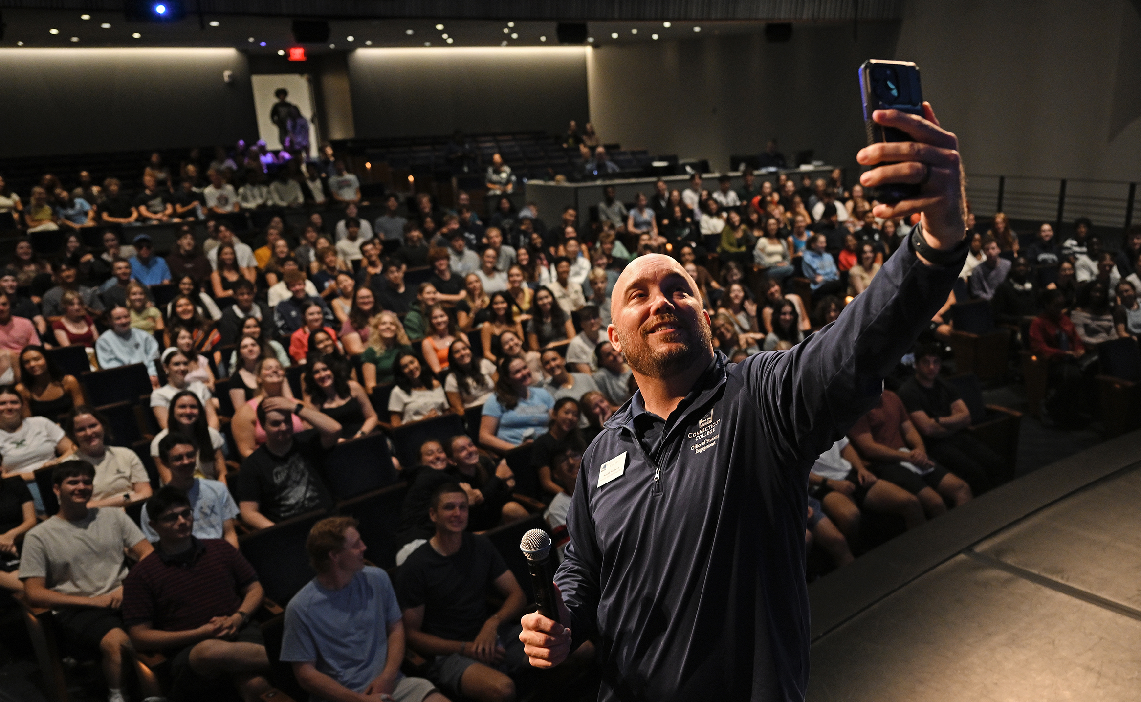 A college dean takes a selfie photo with an auditorium full of first year students.