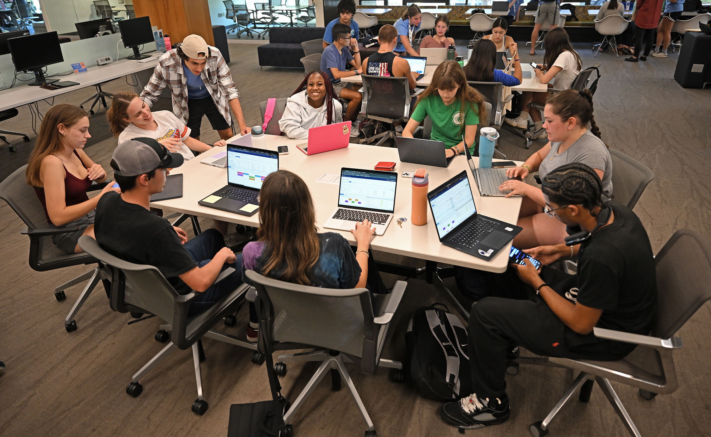College students gather around a table with their laptop computers to complete their course registration in a college library setting.