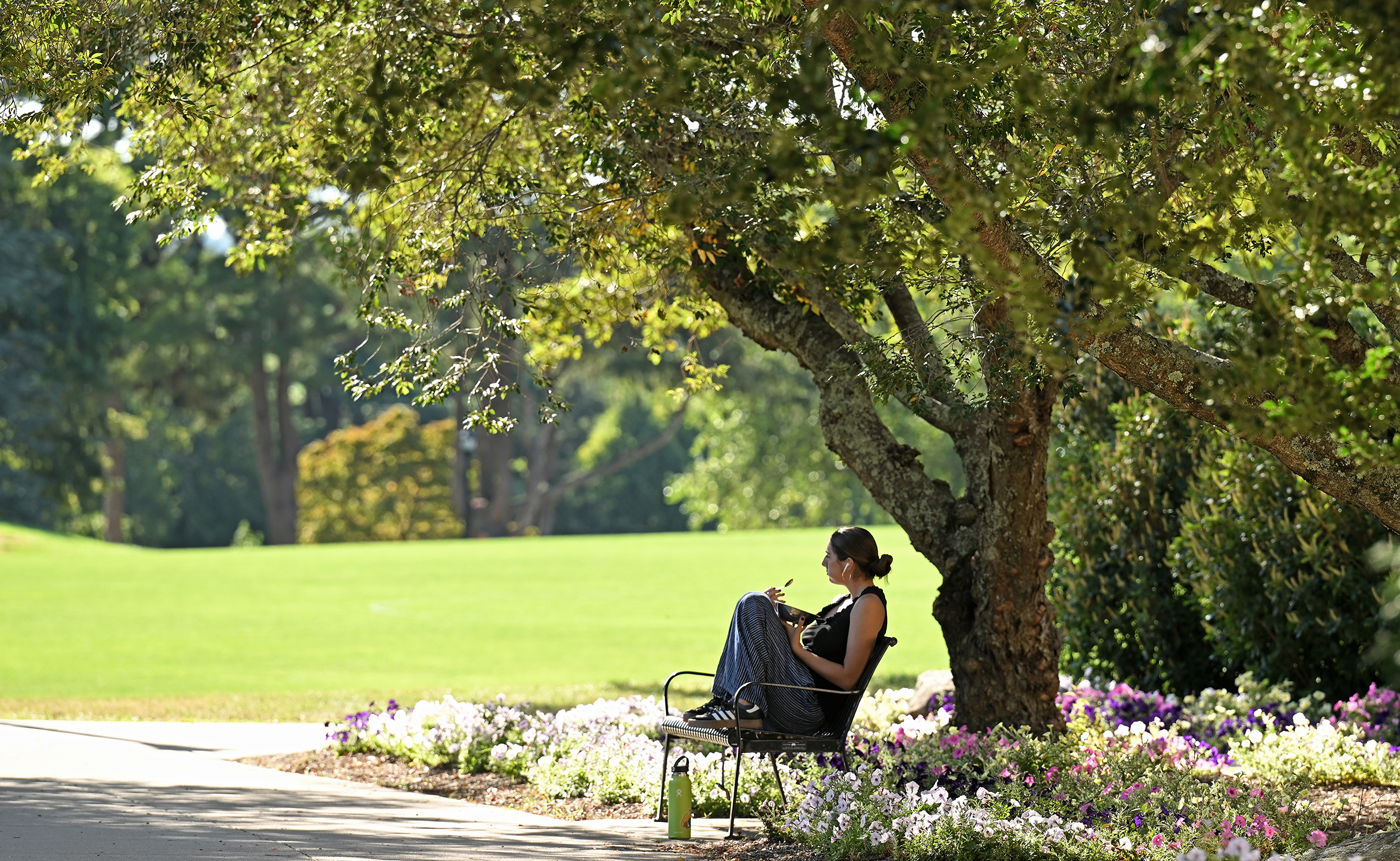 A college student eats breakfast on a shaded bench surrounded by trees and flowers.