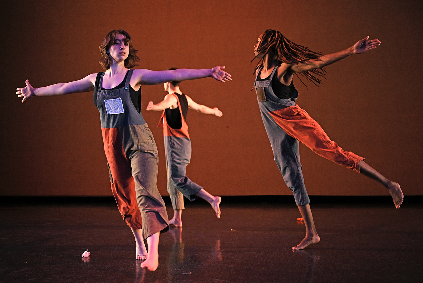 A trio of dance students wearing orange, blue, and gray jumpsuits perform a modern dance piece.