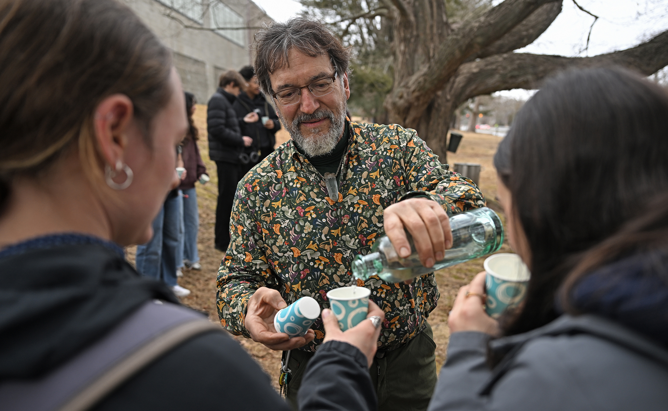 A bearded college professor pours raw maple sap into cups for students to sample.