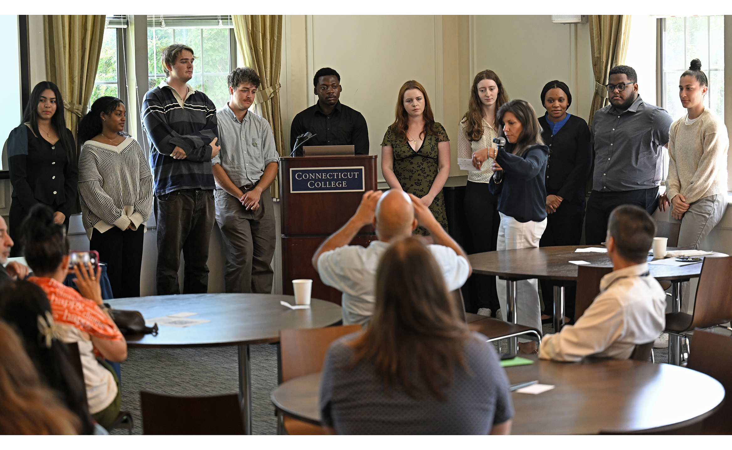 A group of college students stand at the front of a room to give final presentations from their summer internships.