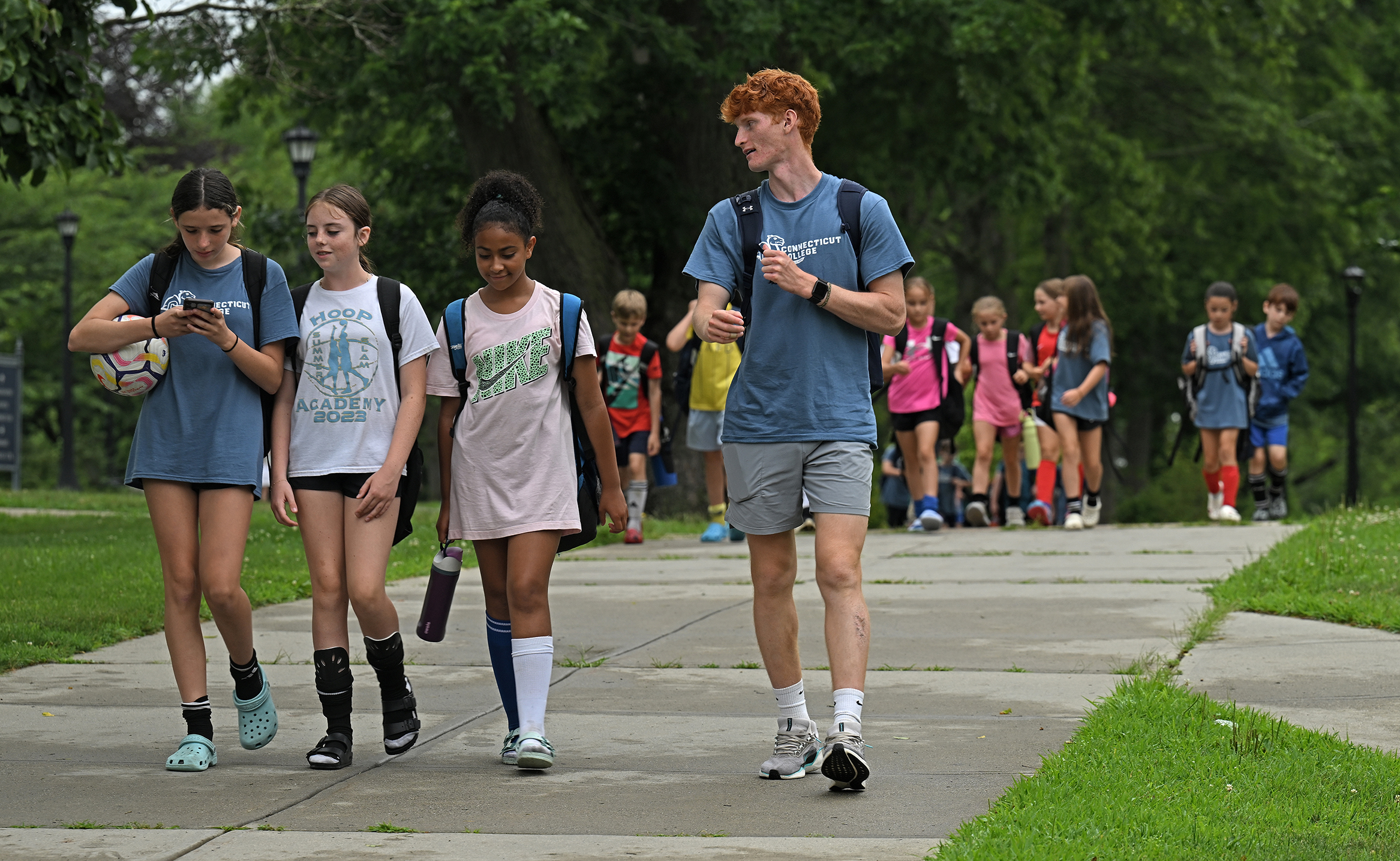 A college soccer player walks and talks with middle school students walking along a concrete path during a summer soccer camp program.