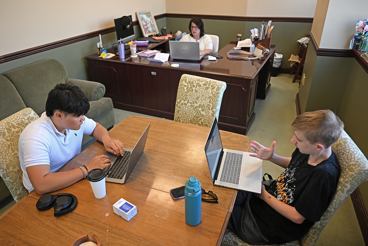 Two students with laptop computers sit at a table and consult with the supervisor at a nearby desk during a summer internship.
