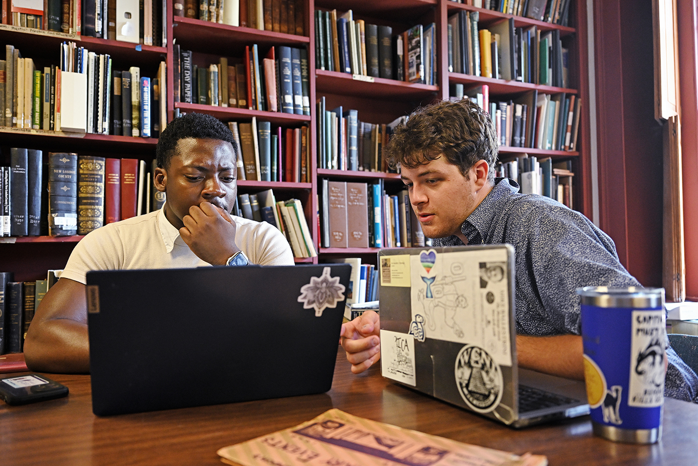 Two students gather around a laptop computer in front of a wall of books during a summer internship.