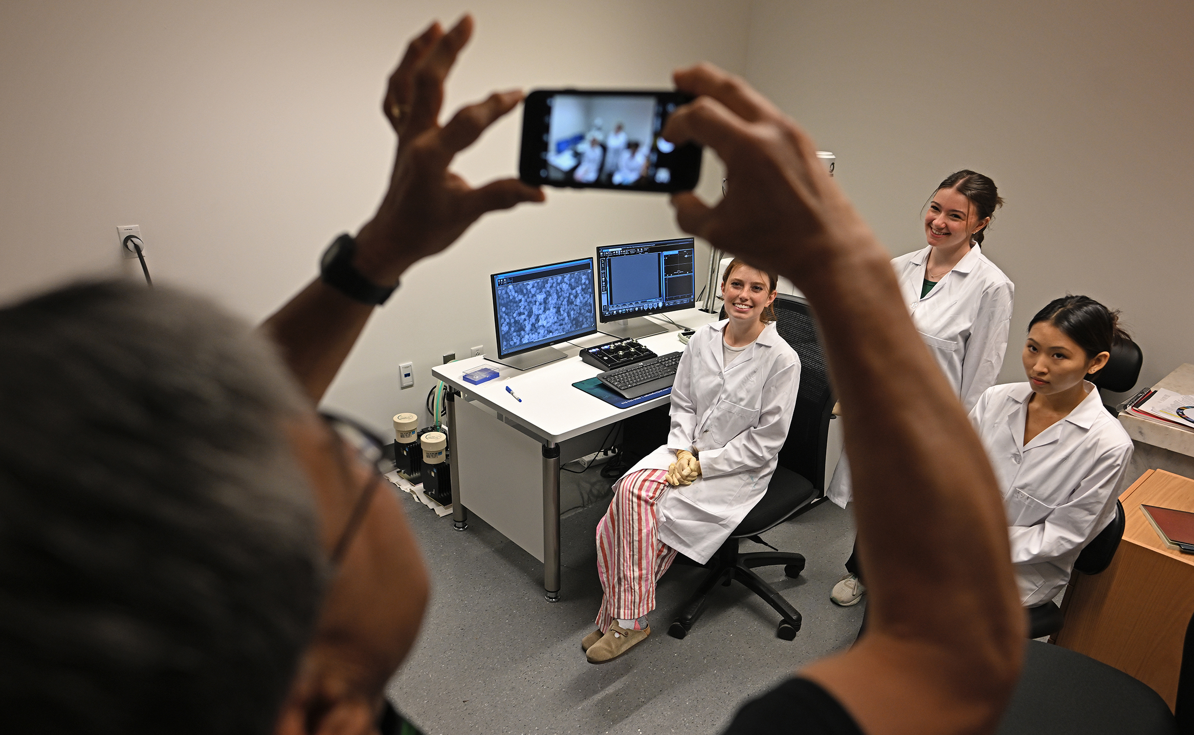 A professor takes a photo of three student researchers in lab coats posing with an electron microscope.
