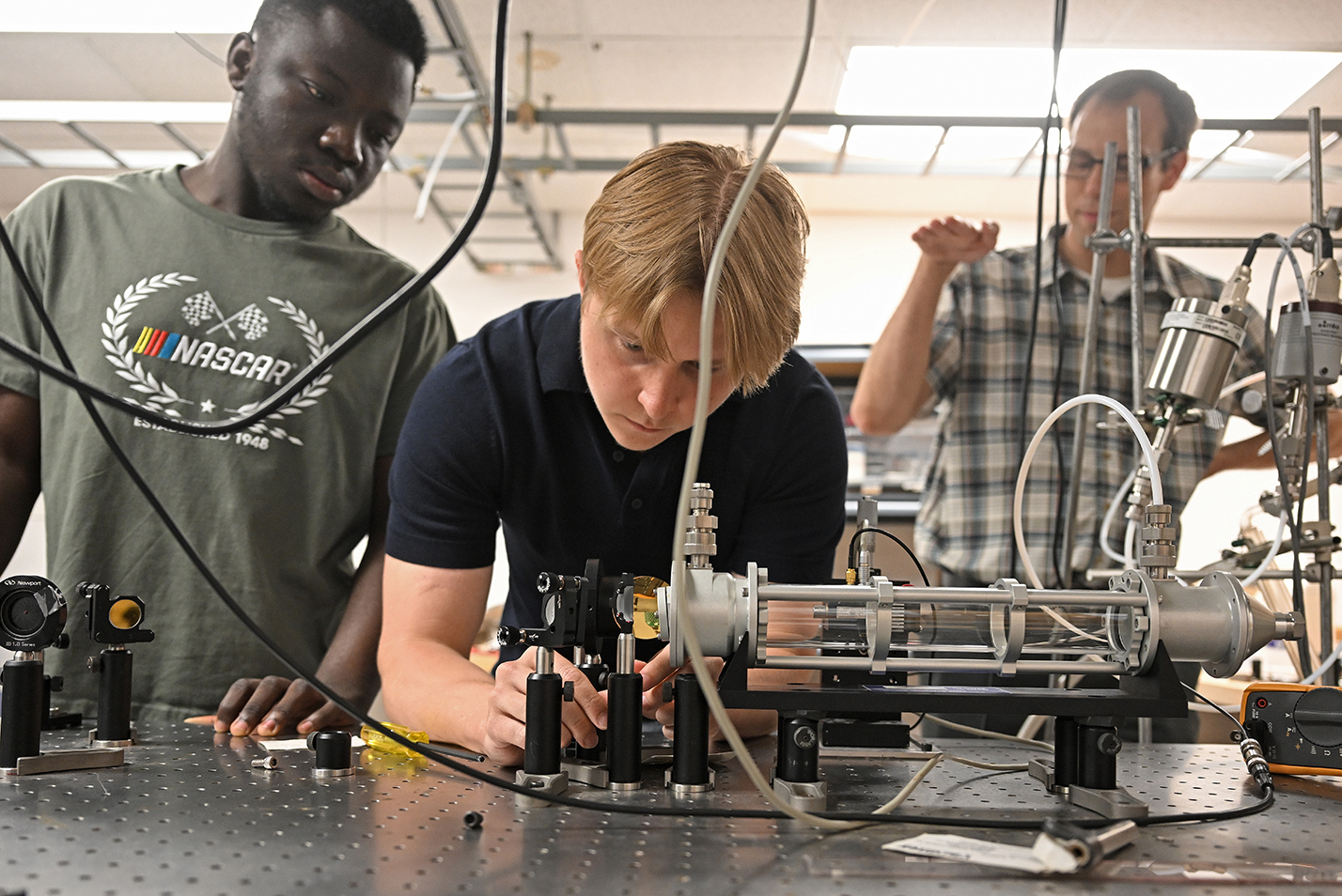 Two student researchers work on a laser setup as a professor gives instruction in a research lab.