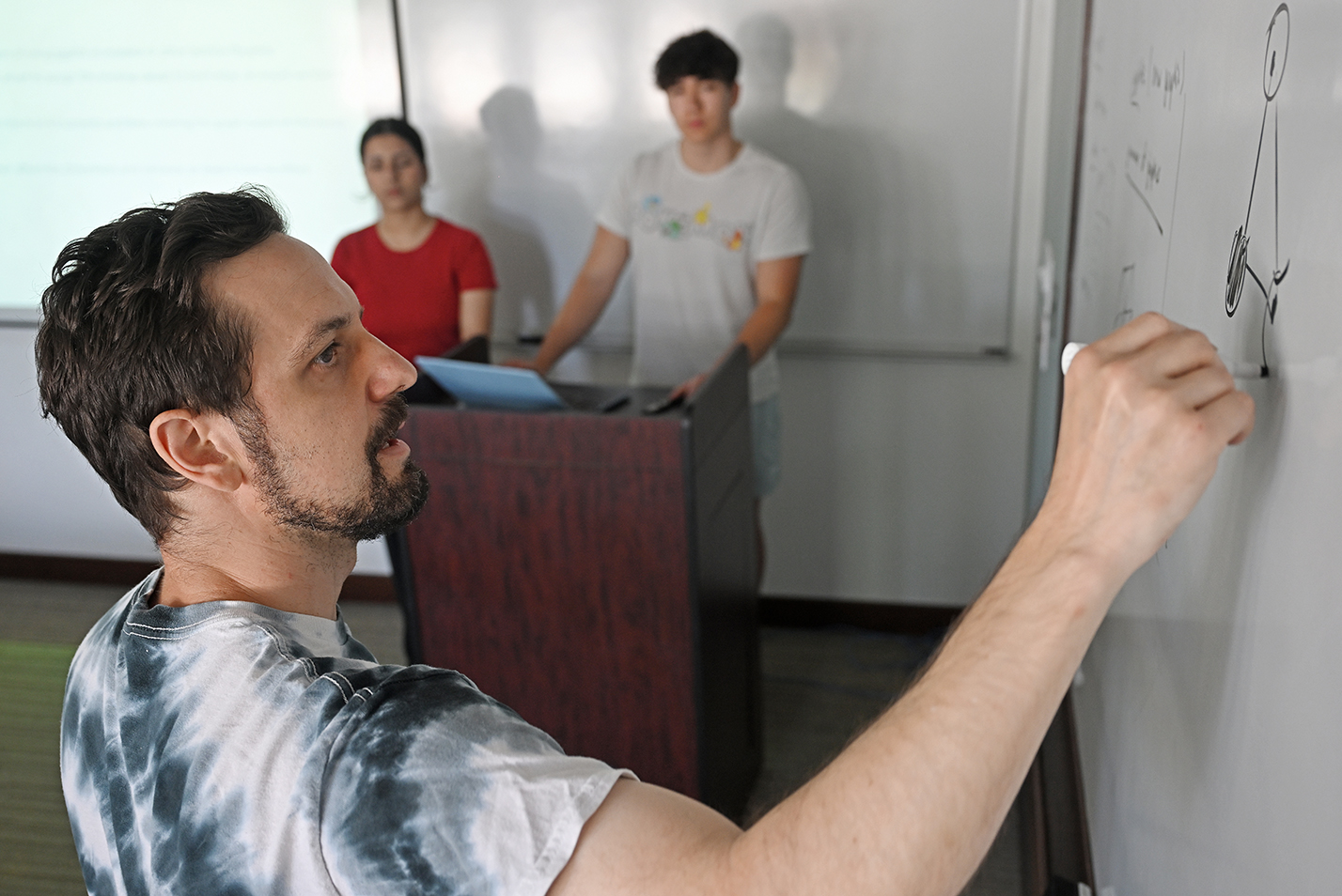 A professor writes equations on a white board for two students standing at a podium.