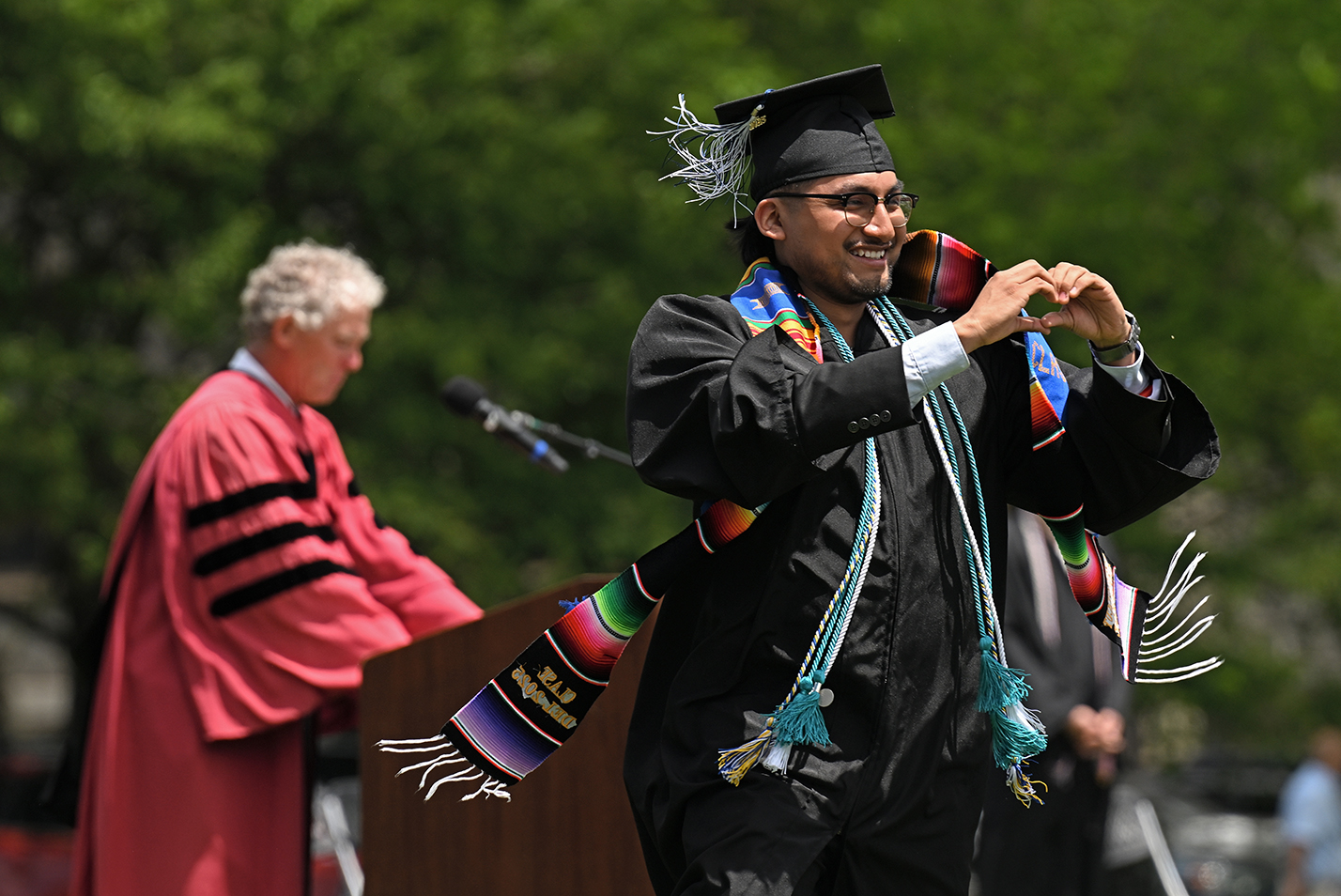 A student in graduation regalia gestures toward the audience as he walks across the stage to receive his diploma.