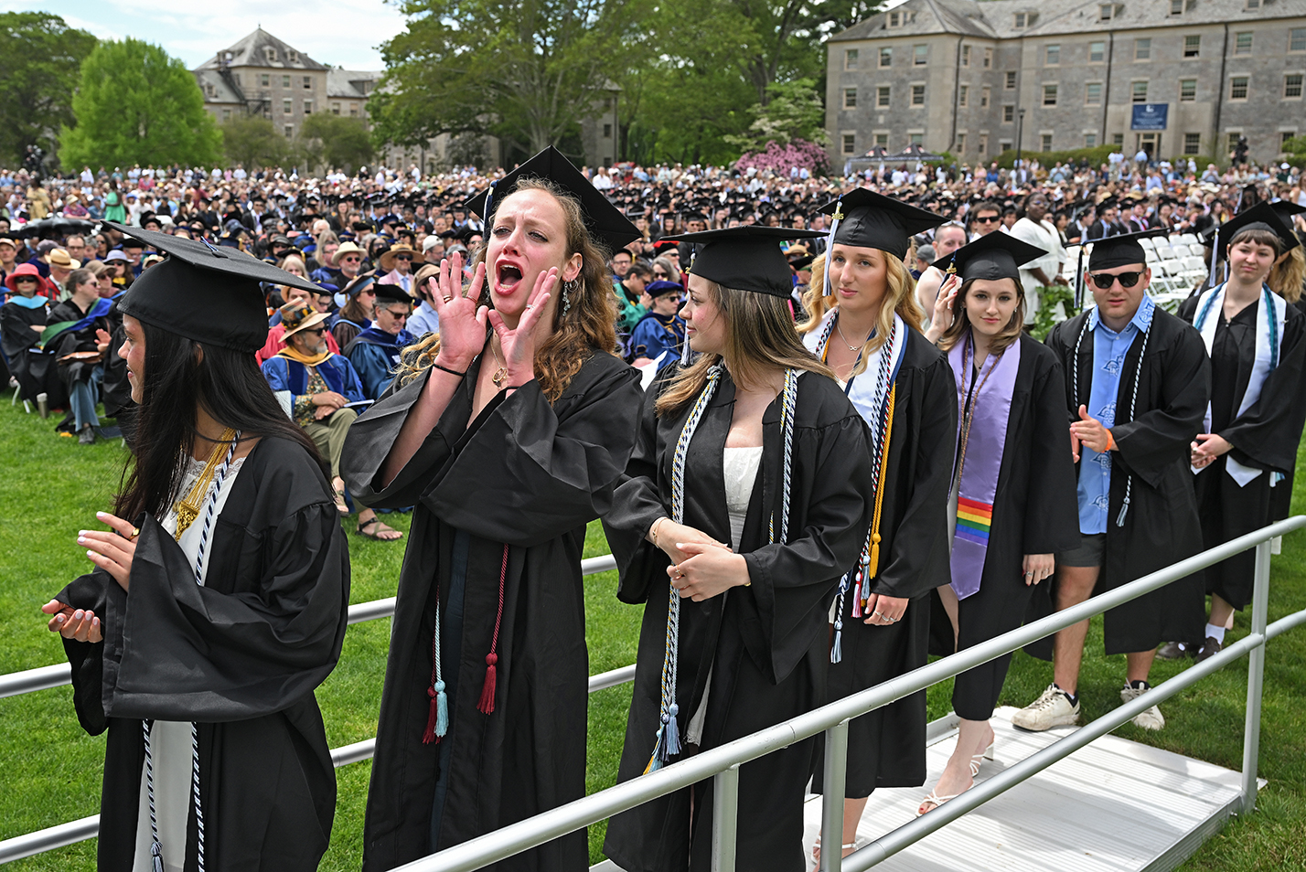 One graduate in the line holds their hands to their mouth and shouts encouragement to fellow graduates during commencement exercises.