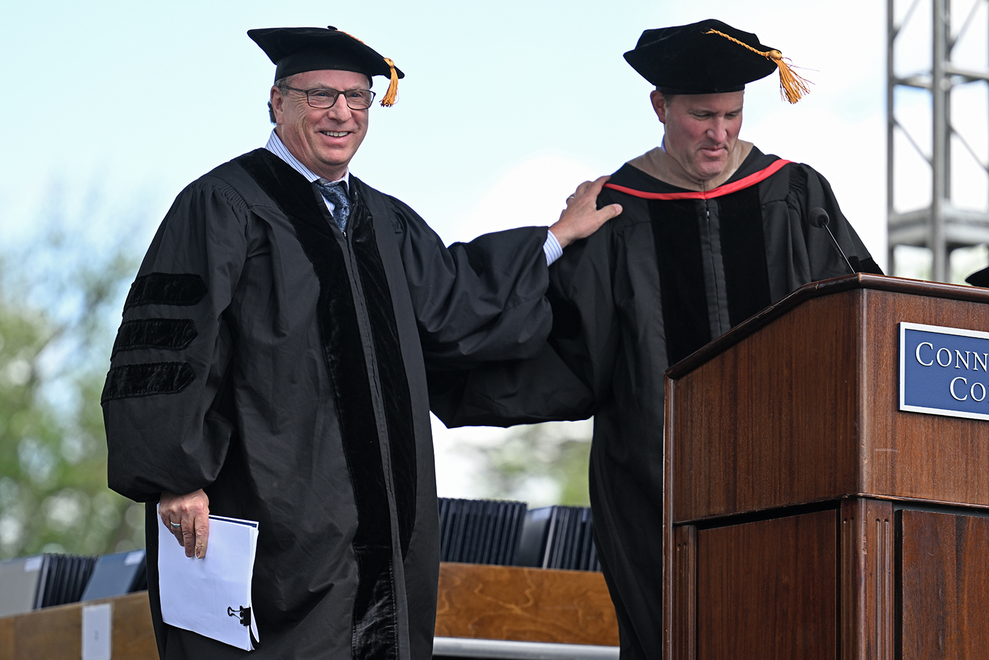 Two academics in regalia embrace on the stage during commencement exercises.