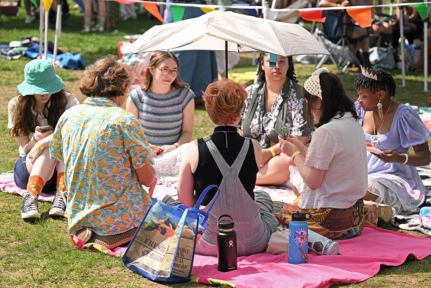 A group of students gather and play cards on a blanket between performances at an outdoor music festival.