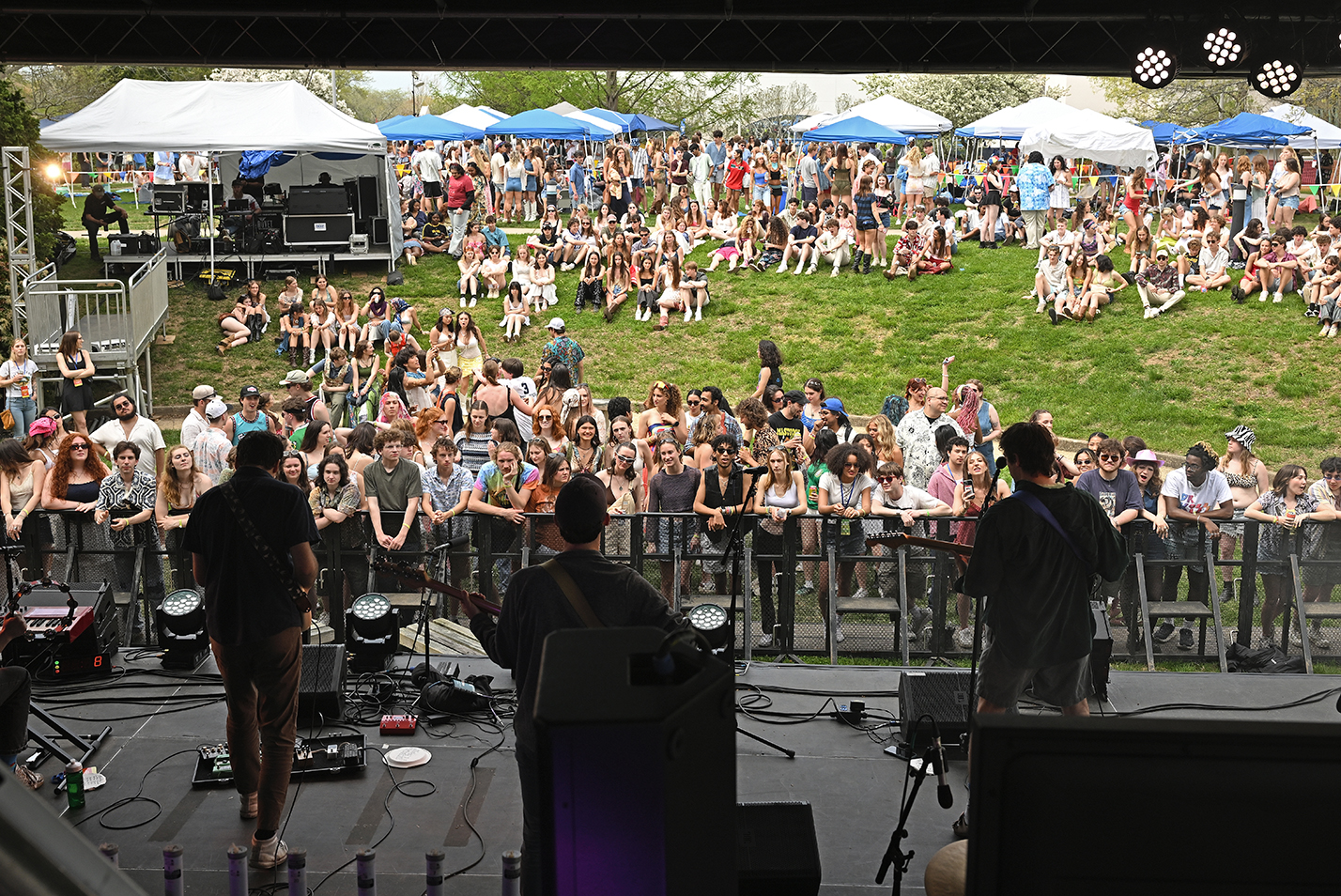A student band performs on stage as the audience gathers on the grassy hill to watch during an outdoor music festival.