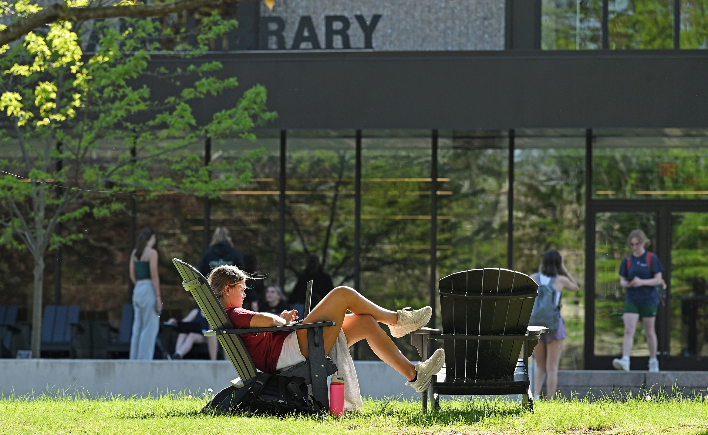 A student sits in one Adirondack chair with her feet up on another and works on a laptop computer on a sunny spring day in front of the college library.