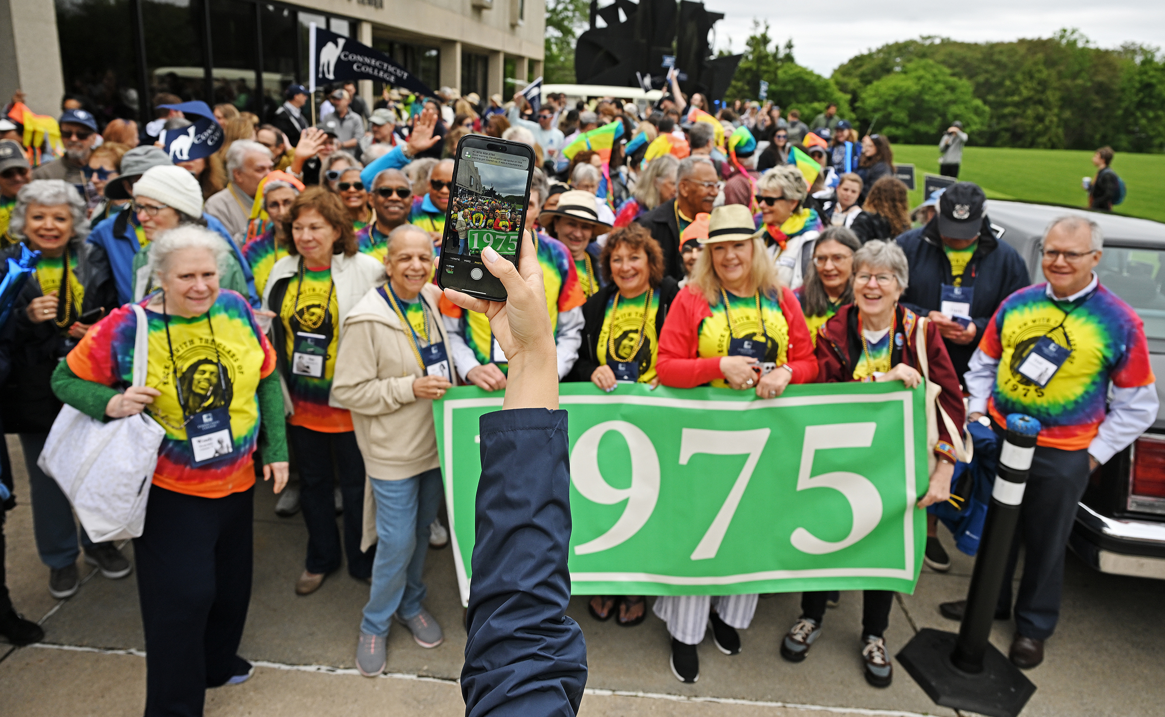 A disembodied hand holds a smart phone high to take a photo of a college class of 1975 reunion group.