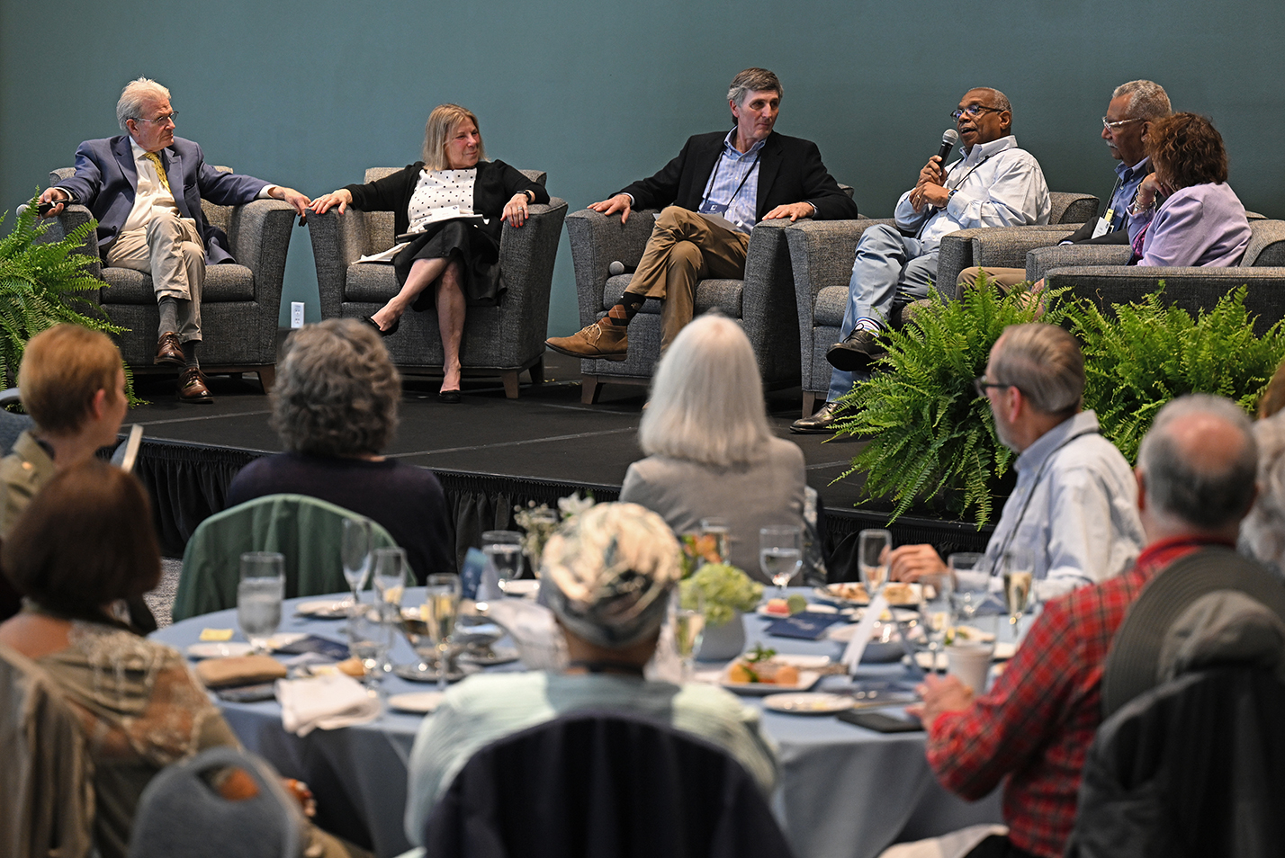 A group of panelists in large chairs sit on stage and hold a discussion in front of an audience.