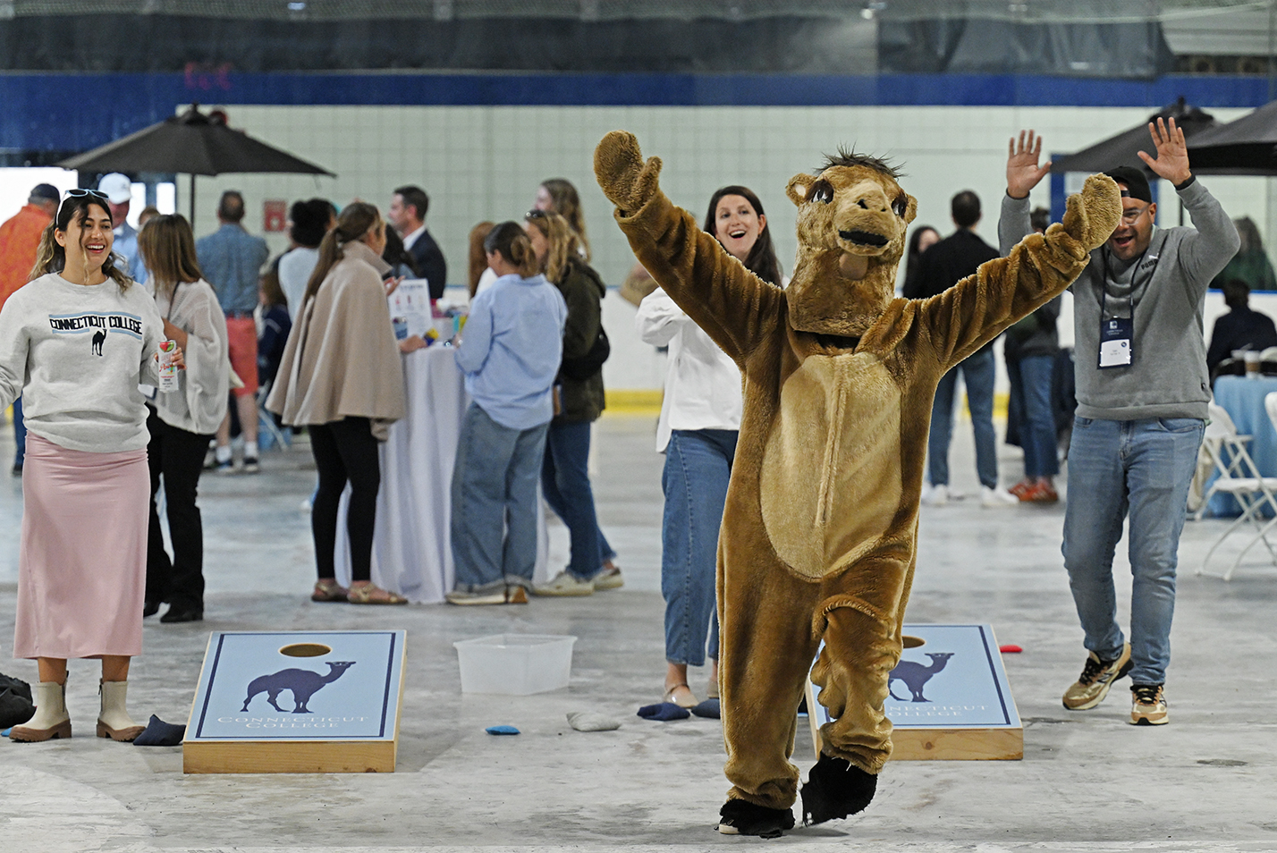 A person in a camel mascot outfit celebrates while playing cornhole with a group of alumni at a reunion event.
