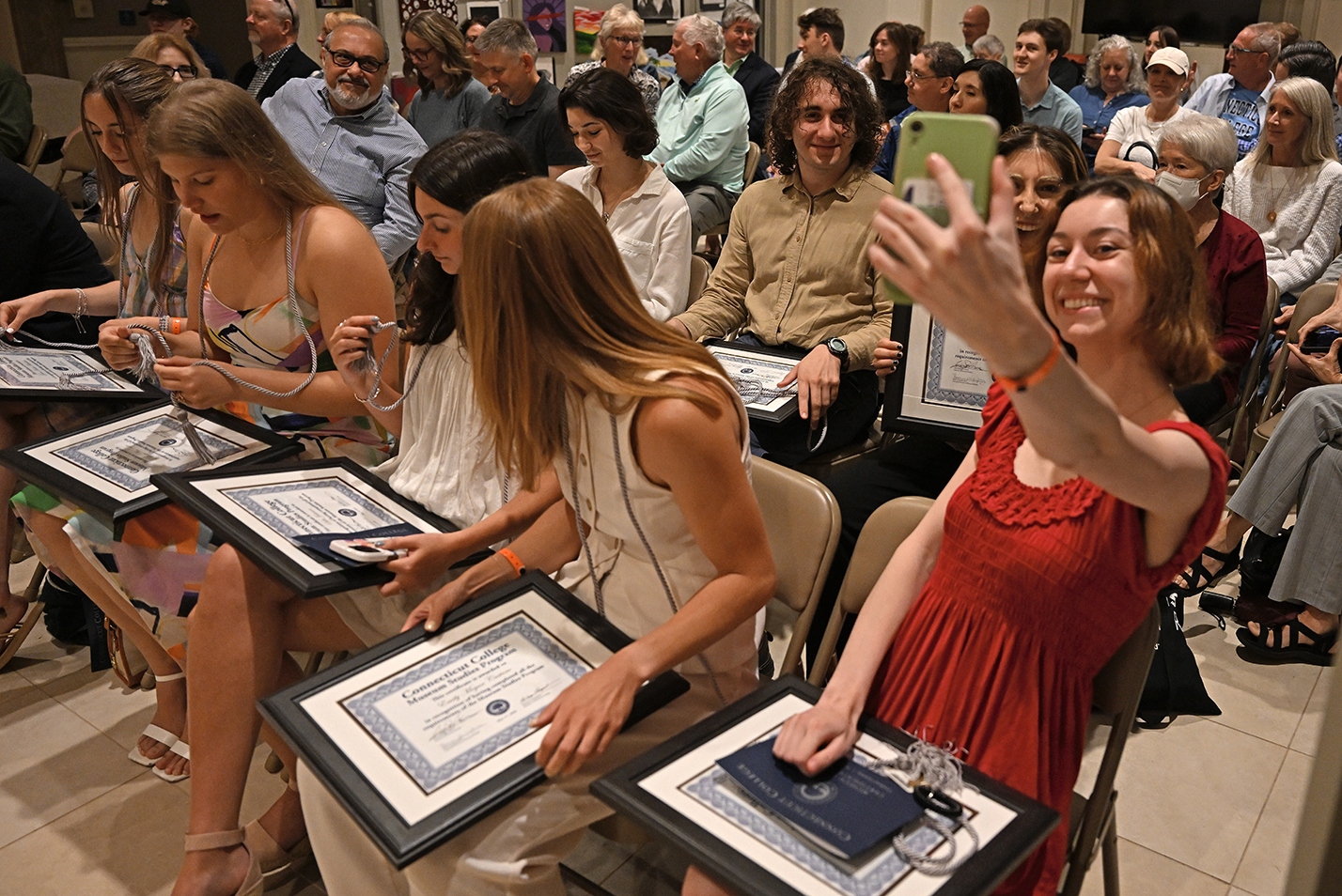 A student in a red dress takes a selfie photo with fellow students at a certificate ceremony.